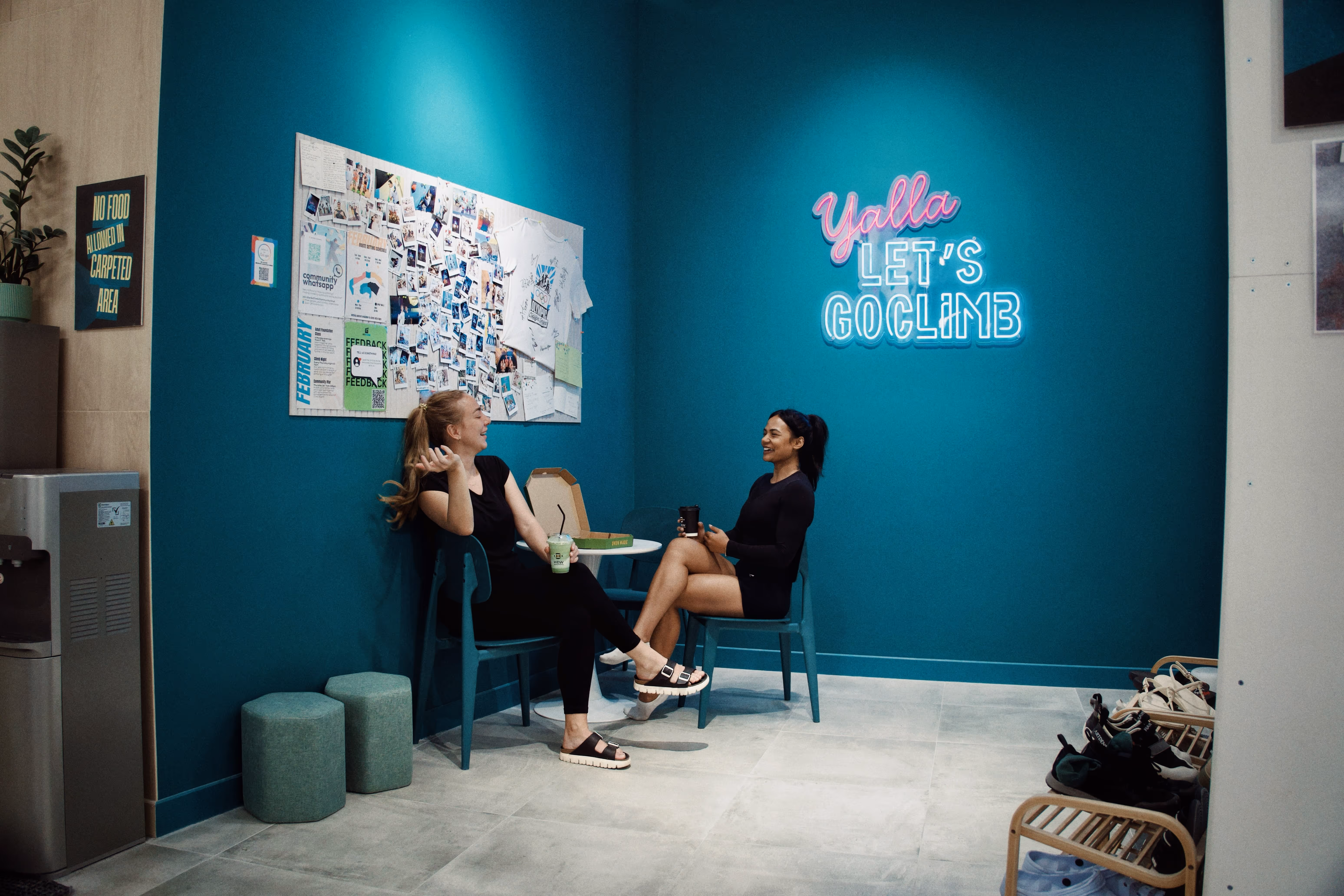Two women sitting and chatting in a teal-colored room with a neon sign reading 'Yalla Let's Go Climb' and bulletin board on the wall.
