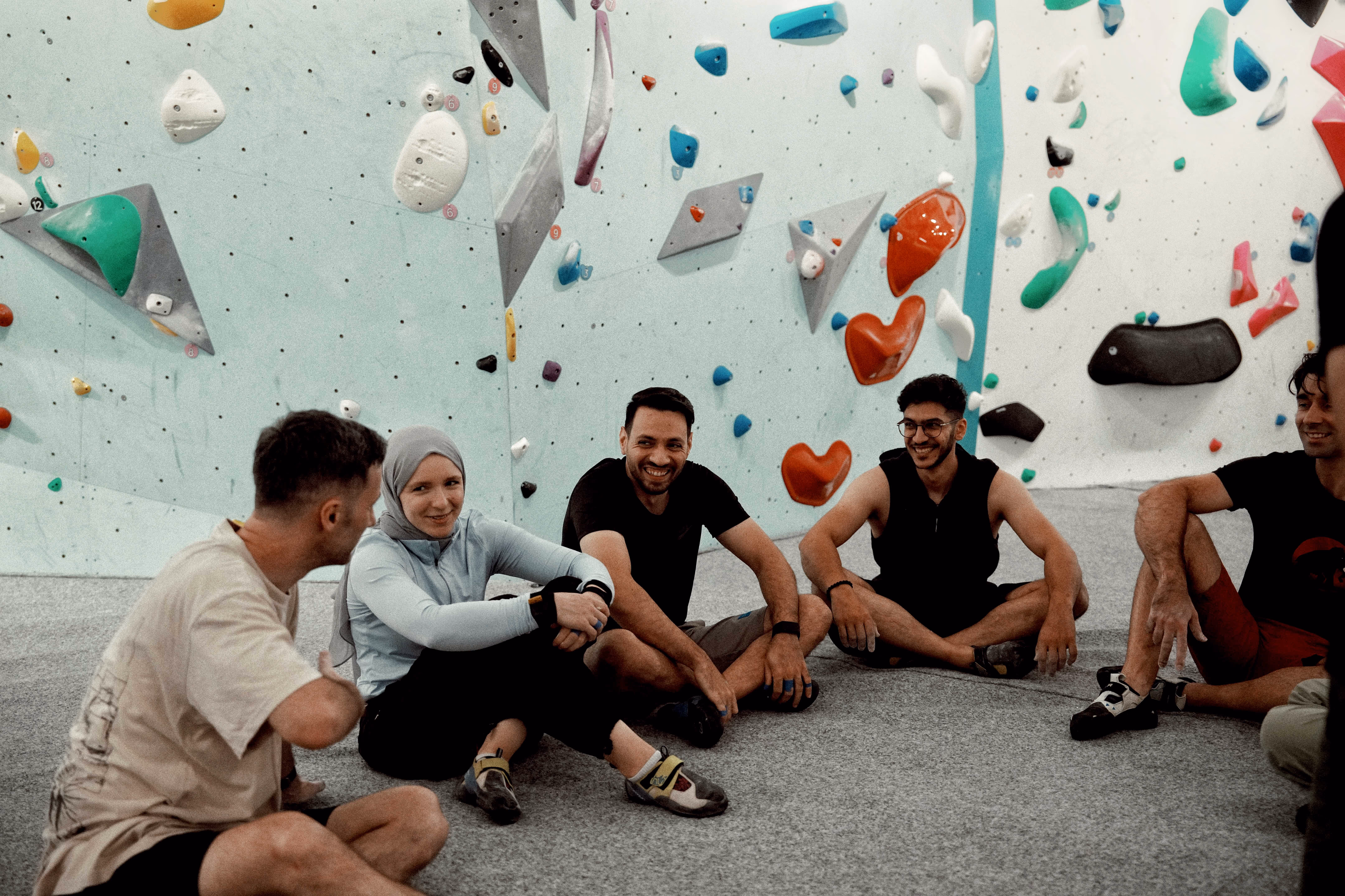 Group of five friends sitting on the floor of an indoor climbing gym, smiling and relaxing near a climbing wall with colorful holds.