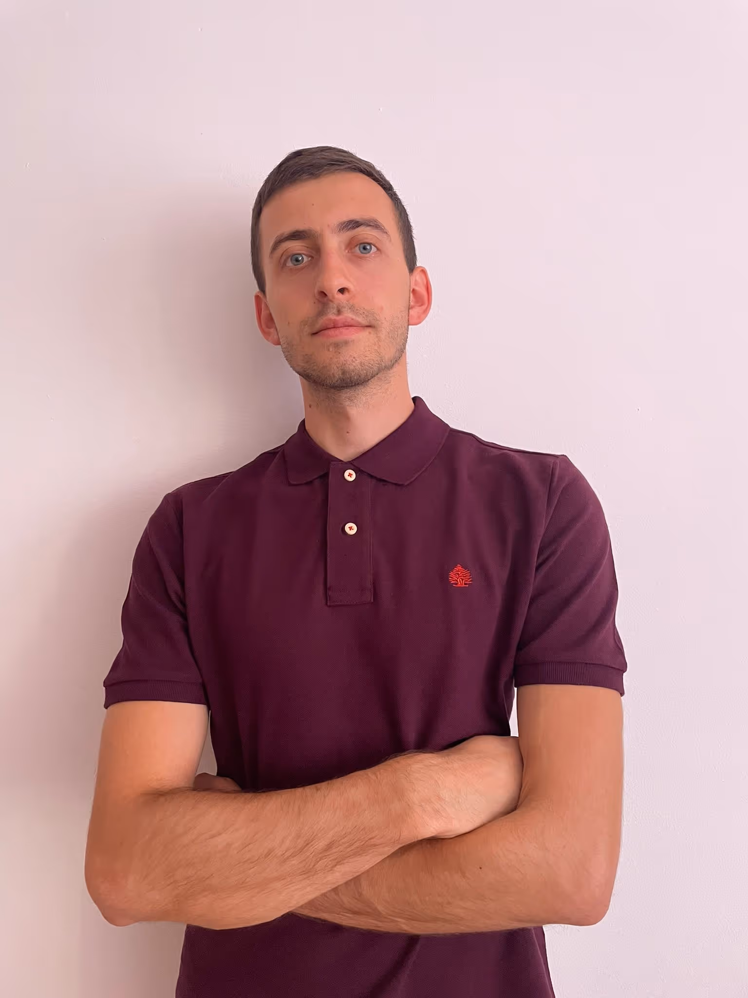 Man with short dark hair and blue eyes wearing a maroon polo shirt with folded arms against a plain light wall.