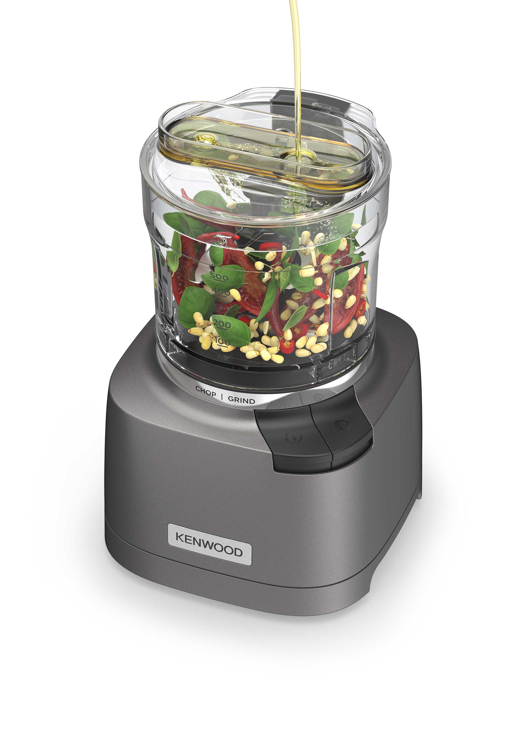Kenwood food chopper with visible ingredients including tomatoes, pine nuts, and greens inside the transparent container, with oil being poured into the top.