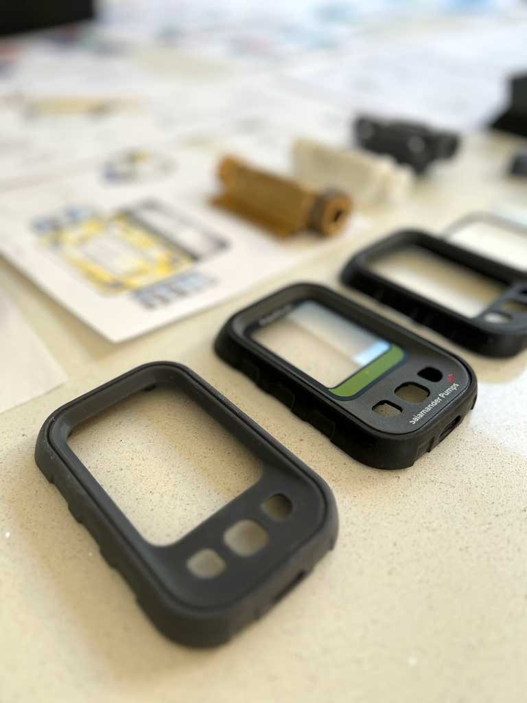 Close-up of four black plastic protective casings for a device, one with a display screen, arranged diagonally on a light surface with blurred sketches and design drawing papers and parts in the background.
