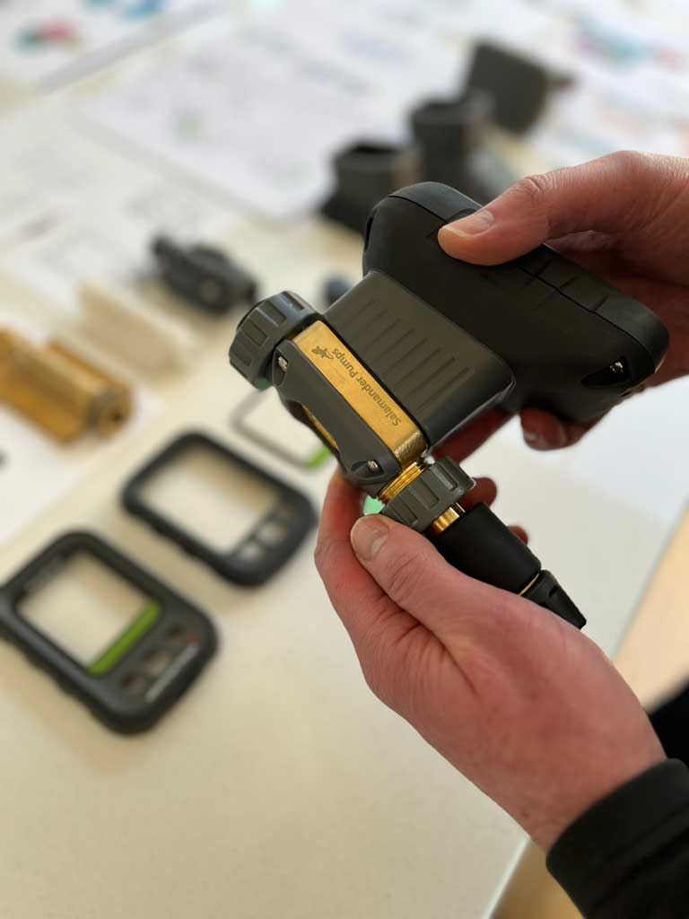 A person holding  the completed Aquascan water meter device, with various electronic and mechanical prototype parts blurred on a table in the background.