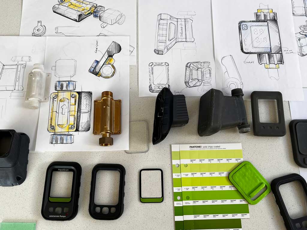 Table with mechanical device sketches, various black and translucent plastic parts, and Pantone solid chips coated color swatches in shades of green.