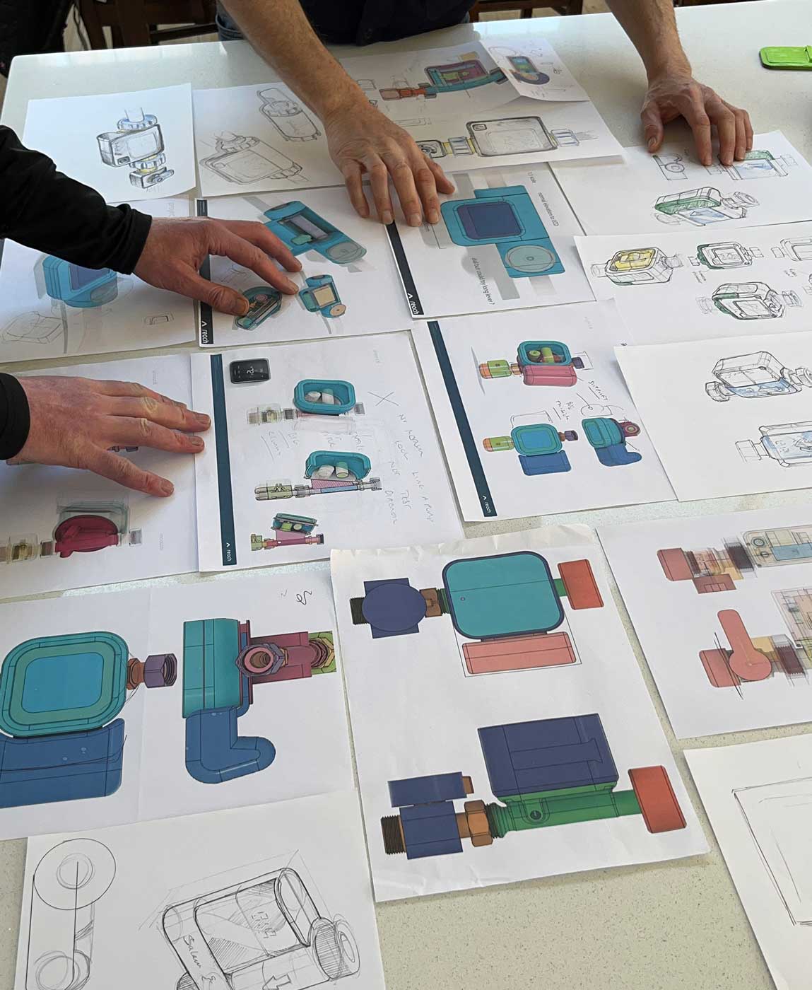 A table covered with multiple colorful technical sketches and 3D renderings of mechanical parts or devices, with two pairs of hands pointing and interacting with the papers.