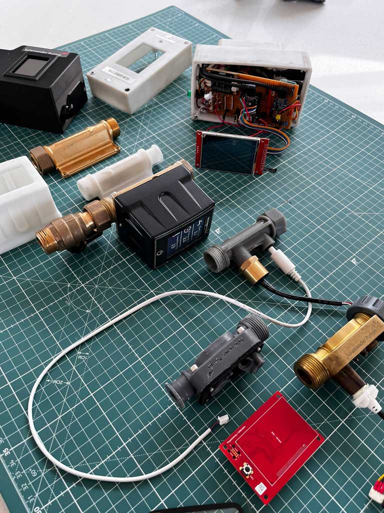 Various electronic components, circuit boards, flow meters, and connectors arranged on a green grid cutting mat, including a Salvamander Pumps labeled brass part and an open electronics enclosure with wiring.