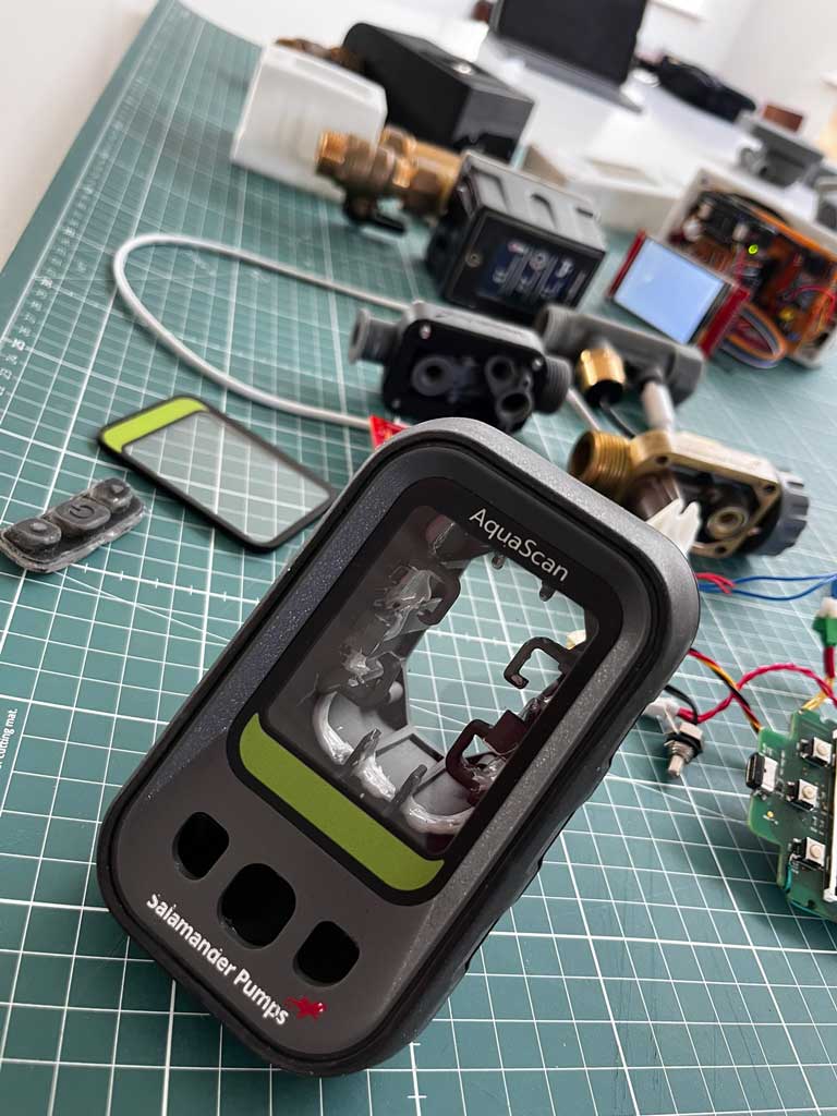 A Salamander Pumps AquaScan device with its casing off, showing internal components, placed on a green grid cutting mat with various other disassembled mechanical and electronic parts in the background.