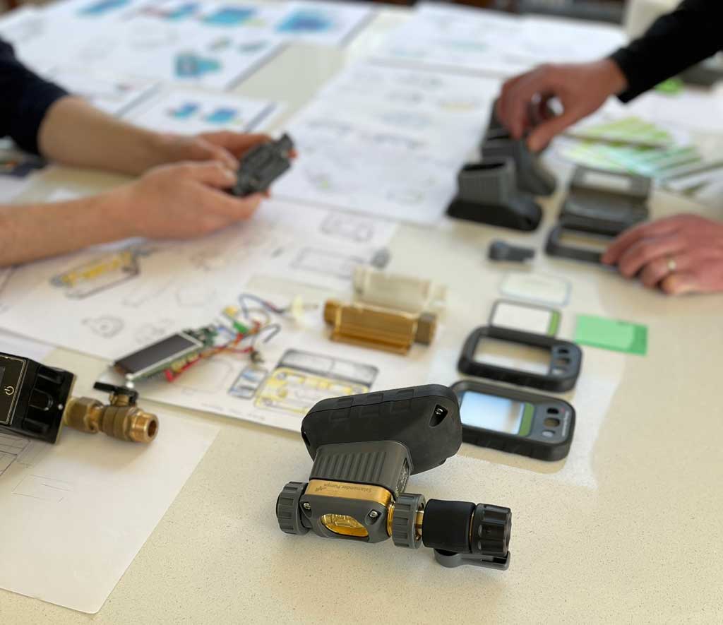 Close-up of hands assembling and examining small mechanical and electronic device parts on a table with technical drawings and multiple device casings.