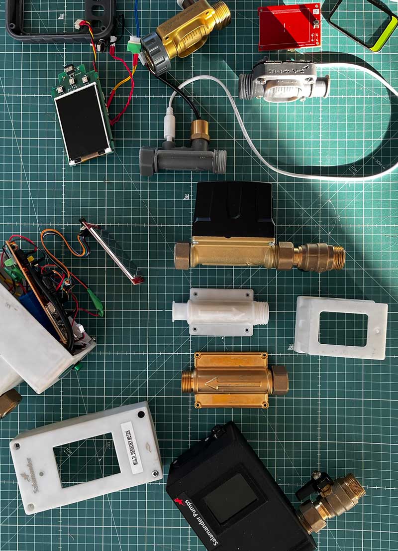 Various water flow sensors, valves, and electronic parts laid out on a green grid cutting mat.