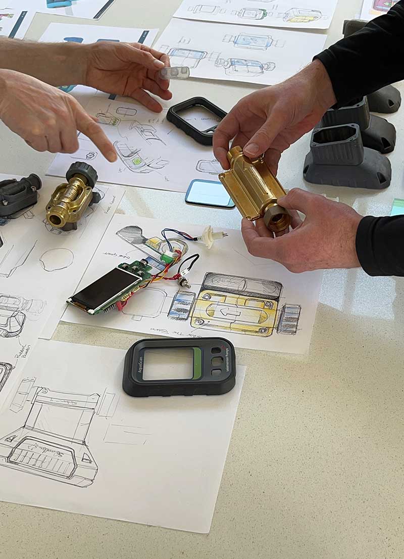 Two people examining a brass mechanical part over sketches and electronic components on a table.