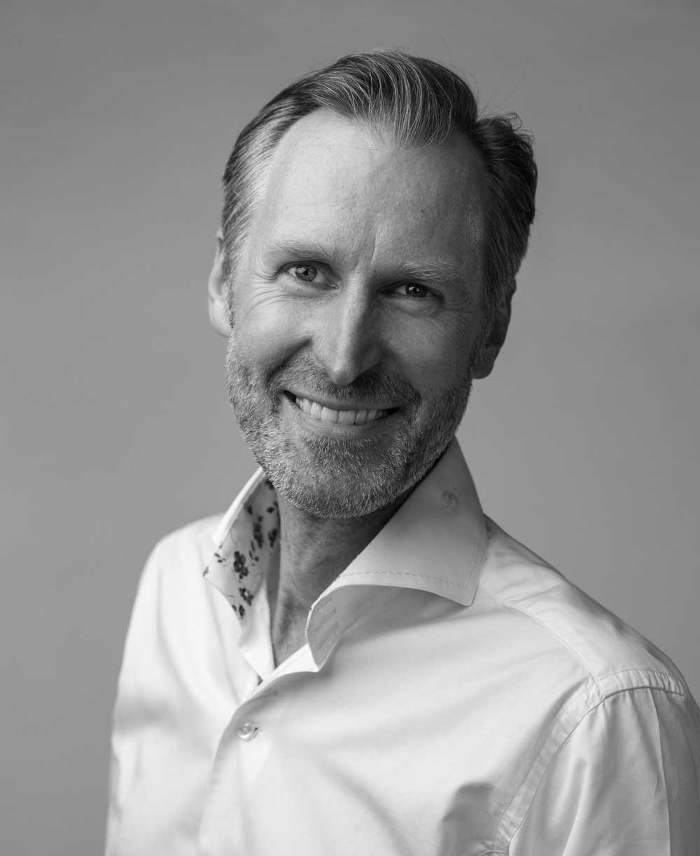Black and white portrait of a middle-aged man with short hair and a beard, smiling warmly and wearing a white collared shirt with a patterned inner collar.