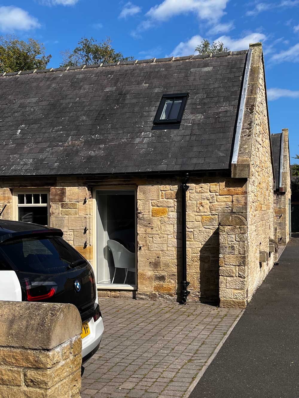 Stone barn house with a steep dark slate roof, a small window on the roof, a glass door showing a modern white chair inside, and a black and white BMW car parked on the cobblestone driveway.