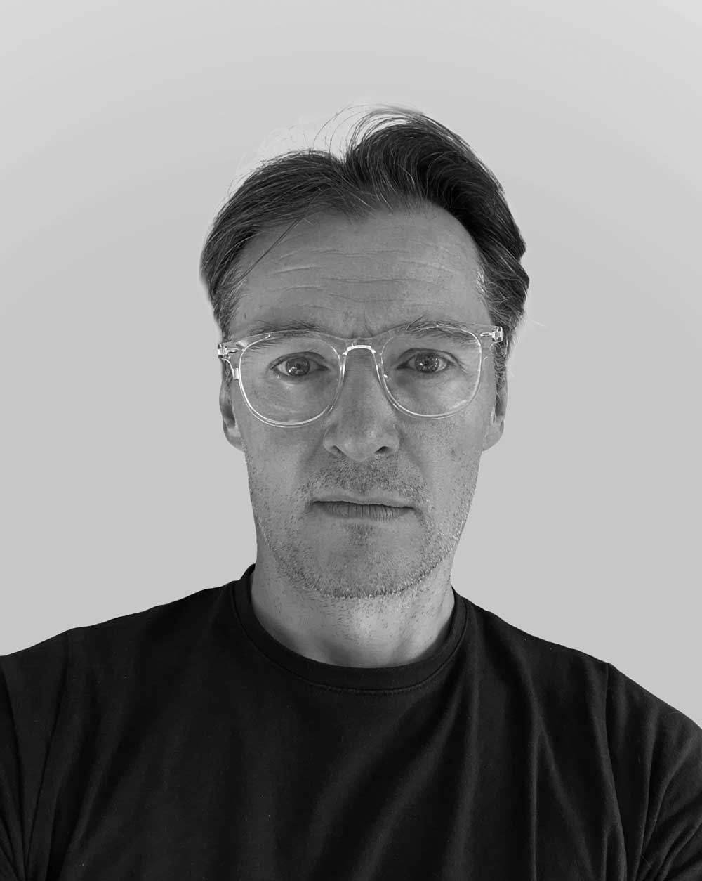 Black and white close-up portrait of a man with short hair, clear-framed glasses, and a neutral expression wearing a dark shirt.