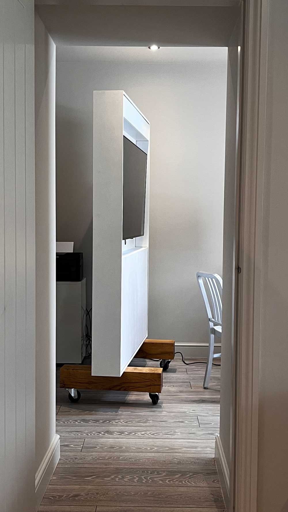 Side view of a modern white conference monitor partition on 2 oak wooden blocks sat on casters in a room with grey wood flooring and a metal chair on the right.
