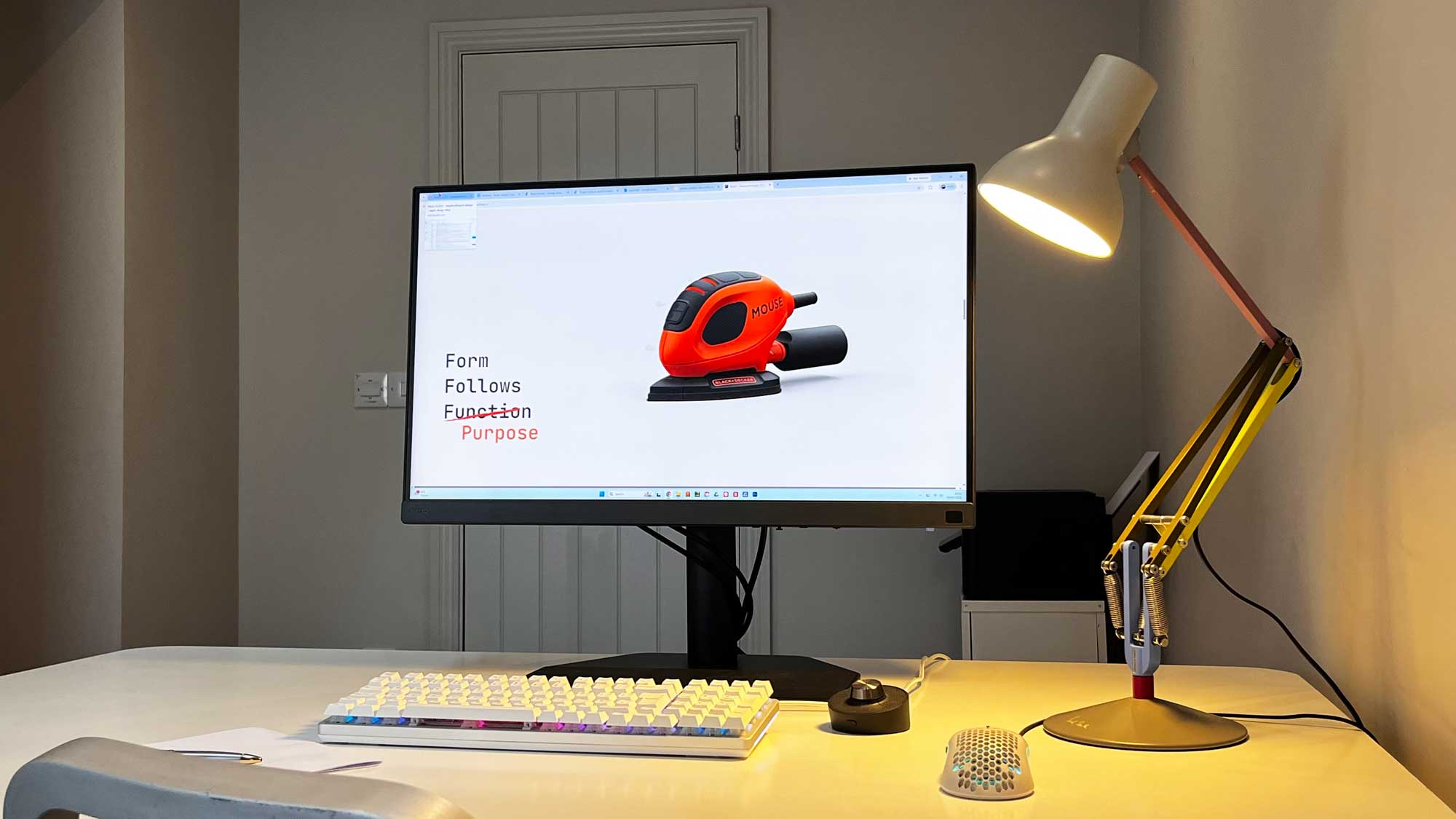 Modern workspace with a large monitor displaying an image of an orange handheld tool labeled 'MOUSE' with text reading 'Form Follows Function' with 'Function' crossed out and replaced by 'Purpose', a colorful mechanical keyboard, a white honeycomb-patterned mouse, a small control knob, and a yellow adjustable desk lamp on a clean white desk.
