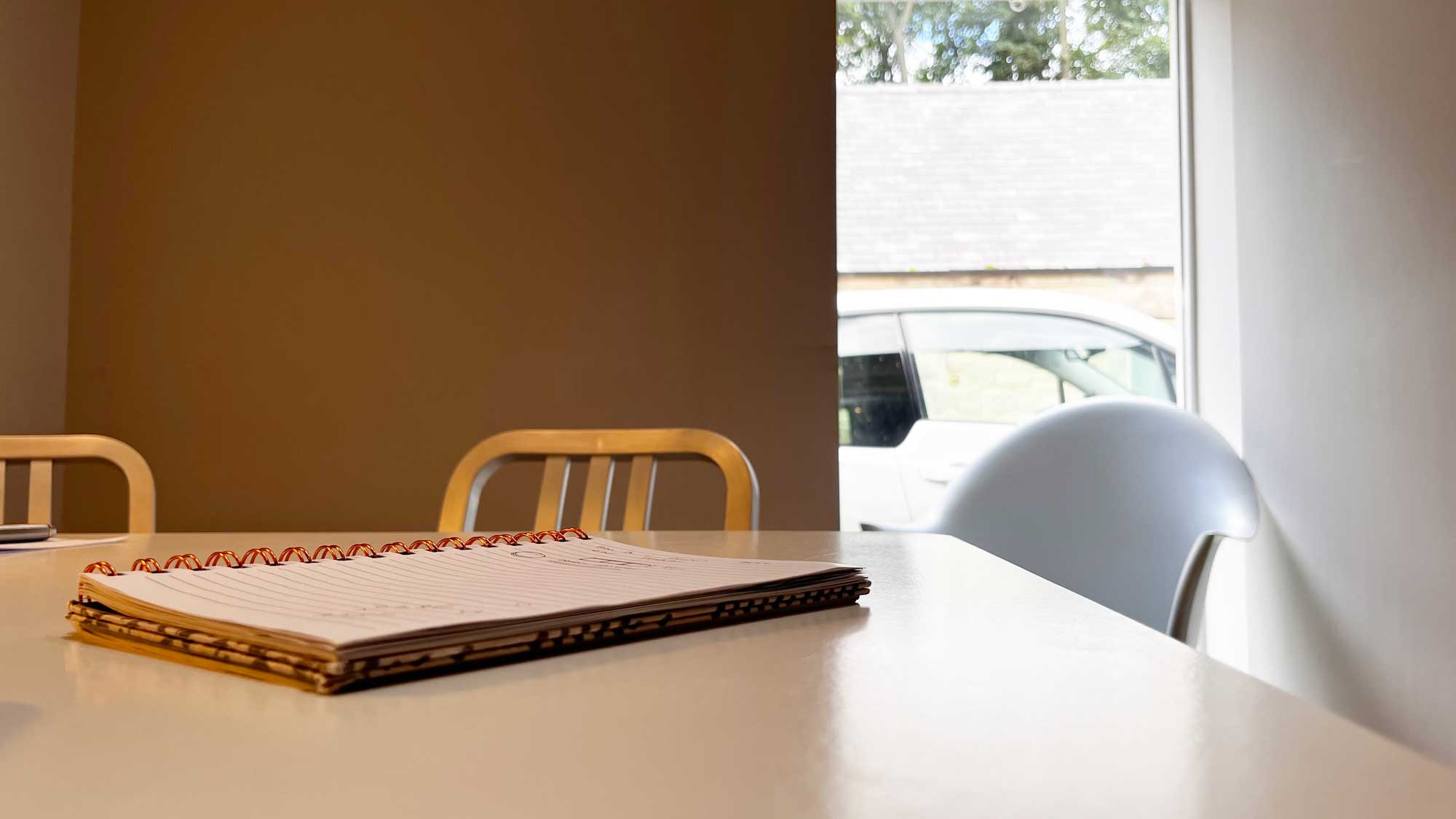 Spiral notebook with lined paper on a white table in a meeting room, with metal chairs and a window showing a parked white car outside.