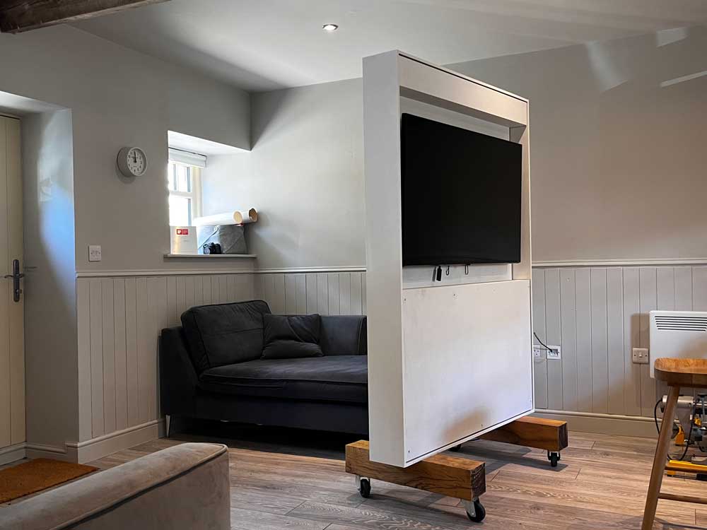 Modern room with a navy blue sofa and a movable white board on wooden block sat upon  trolley wheels that holds a black flat-screen TV facing away from the sofa.