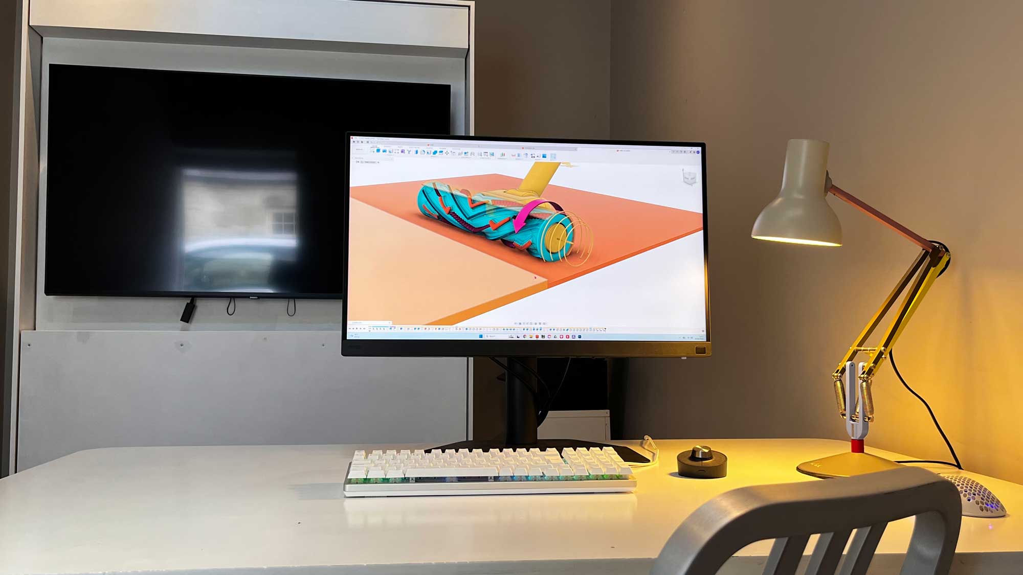 Modern workspace with a white desk, a mechanical keyboard, a single monitor displaying a 3D CAD model with blue and orange elements, a small black device, and a multi coloured angle poise adjustable desk lamp, with an empty chair in the foreground and a large conference monitor screen in the space behind.