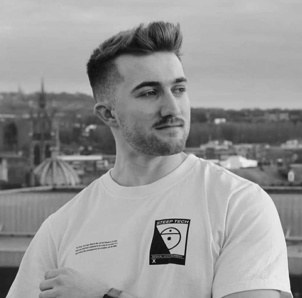 Young man with short hair and a trimmed beard wearing a white T-shirt with a 'Steep Tech' logo, looking to his left with a cityscape background.