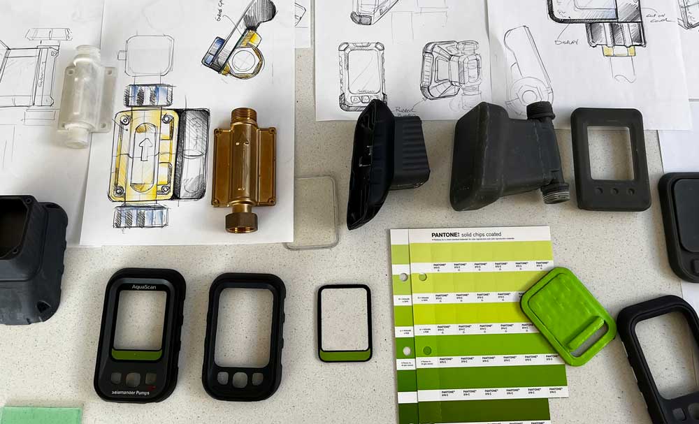 Table with mechanical device sketches, various black and translucent plastic parts, and Pantone solid chips coated color swatches in shades of green.