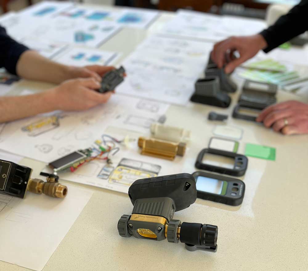 Two pairs of hands evaluating 3D printed black plastic casings for a device, arranged on a light surface with blurred sketches and design drawing papers and parts in the background.