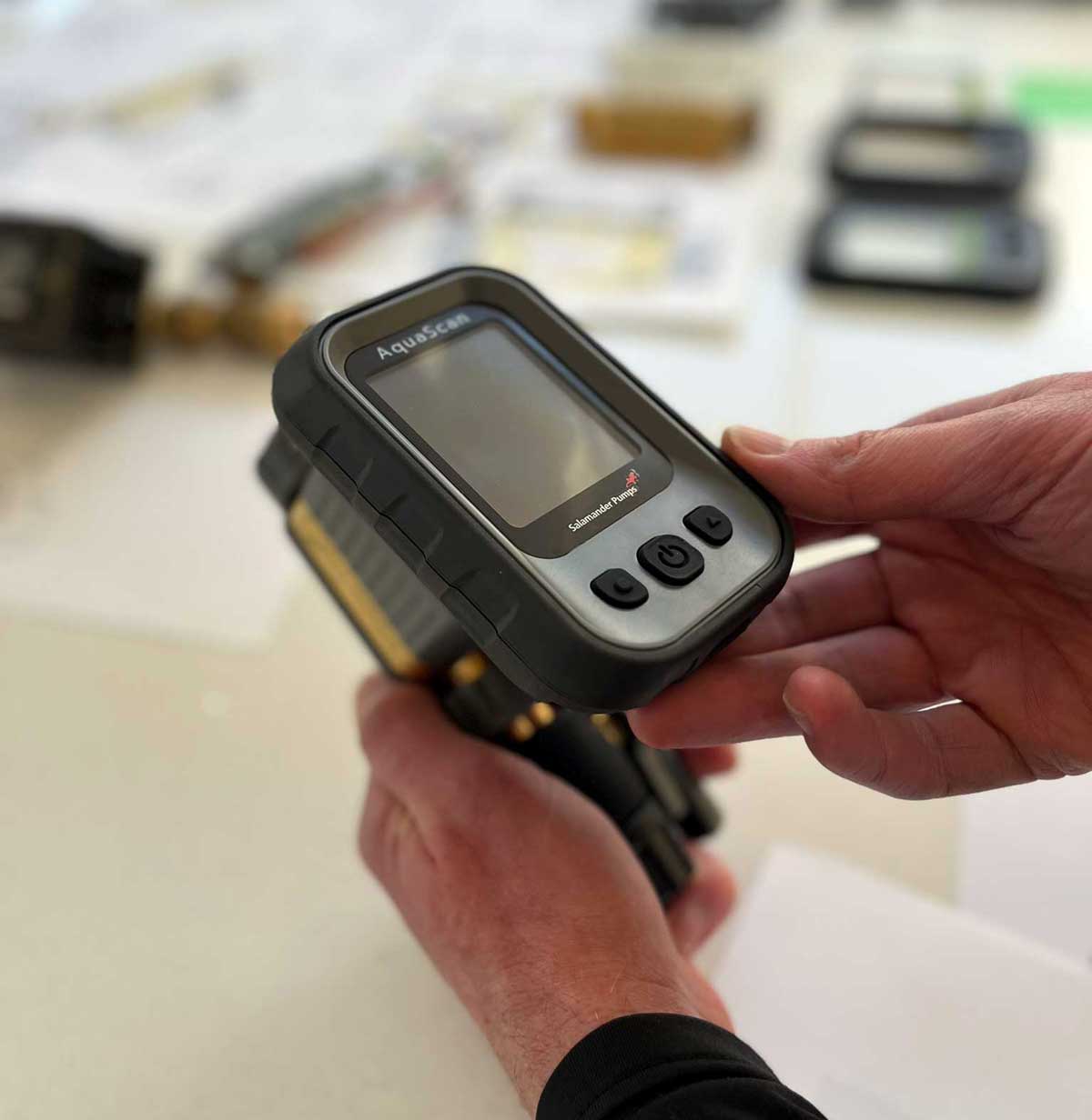 Person holding an AquaScan device with Salamander Pumps branding, featuring a display screen and control buttons on a light-colored surface with blurred objects in the background.
