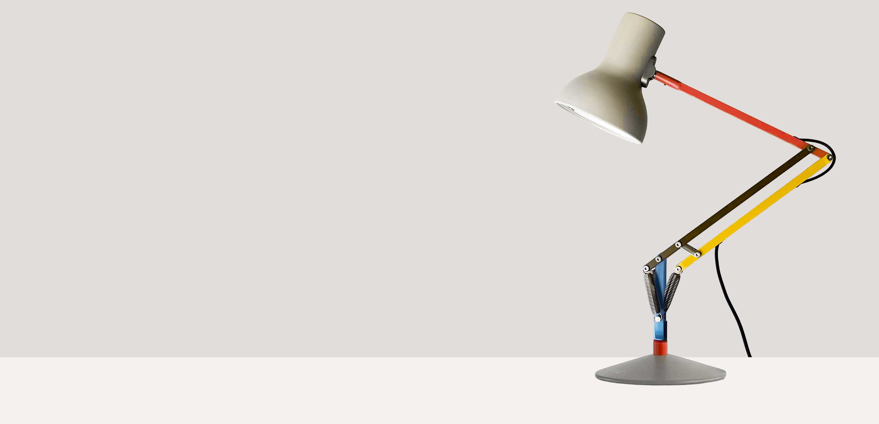 An adjustable desk lamp with a cream lampshade and colourful arms in red, yellow, blue, and metal, sitting on a white table top against a light grey background.