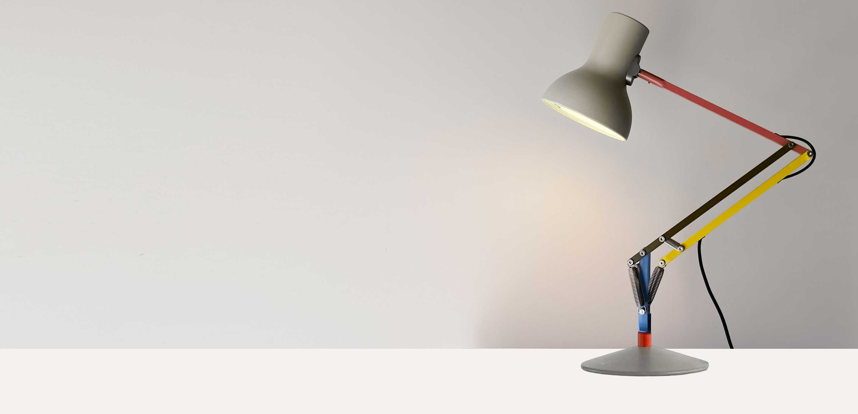 An illuminated adjustable desk lamp with a cream lampshade and colourful arms in red, yellow, blue, and metal, sitting on a white table top against grey background.