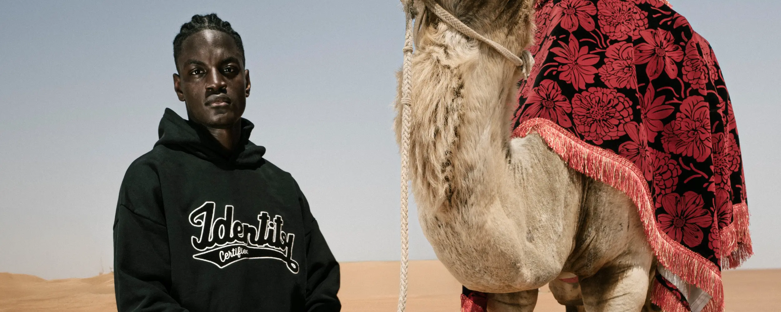 Ibo Ouro-Bodi im schwarzen Certified-Identity-Hoodie neben einem Kamel mit roter Satteldecke in der Wüste.