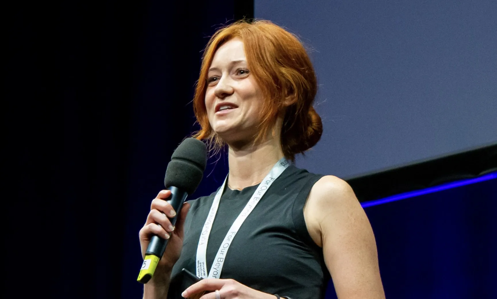 Woman with red hair tied back speaking into a microphone on stage wearing a black sleeveless top and a lanyard.