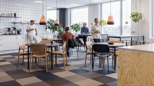 Modern office kitchen and lounge area with several people seated and standing, large windows, hanging plants, and pendant lights.