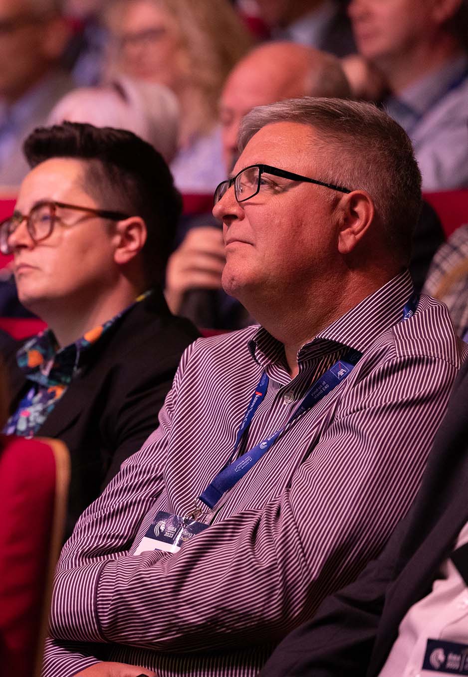 Two attendees sit attentively during the BIBA Conference. The man in the foreground wears glasses, a striped shirt, and a conference lanyard, while the person next to him also wears glasses and business attire.