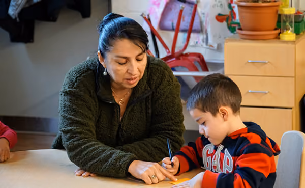 Woman in a green fleece jacket helps a young boy in a red and blue striped hoodie with writing or drawing at a table in a classroom setting.