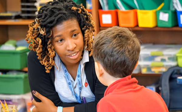 Teacher with curly hair talking attentively with a young student wearing a red shirt in a colorful classroom setting with bins and shelves in the background.