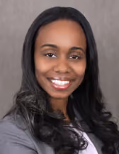 Portrait of a smiling woman with long black hair wearing a gray blazer and a white top against a neutral gray background.
