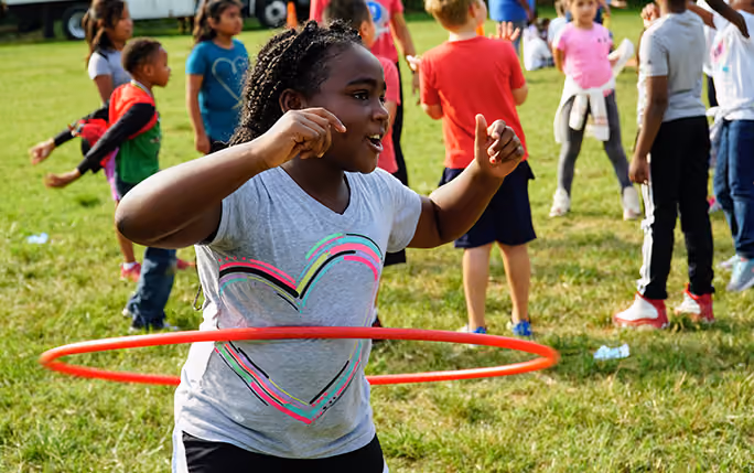 A young girl with braided hair wearing a gray shirt with a colorful heart hula hoops outside on green grass, while other children play in the background.