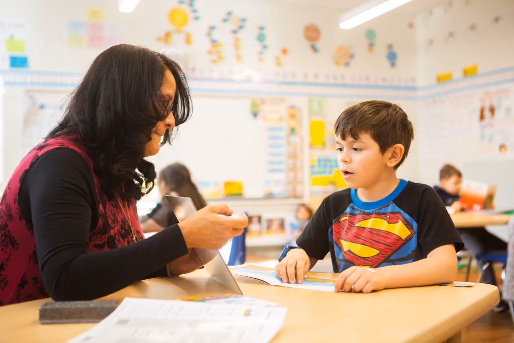Teacher in a classroom engaging with a young boy wearing a Superman t-shirt as they look at a book together at a desk.