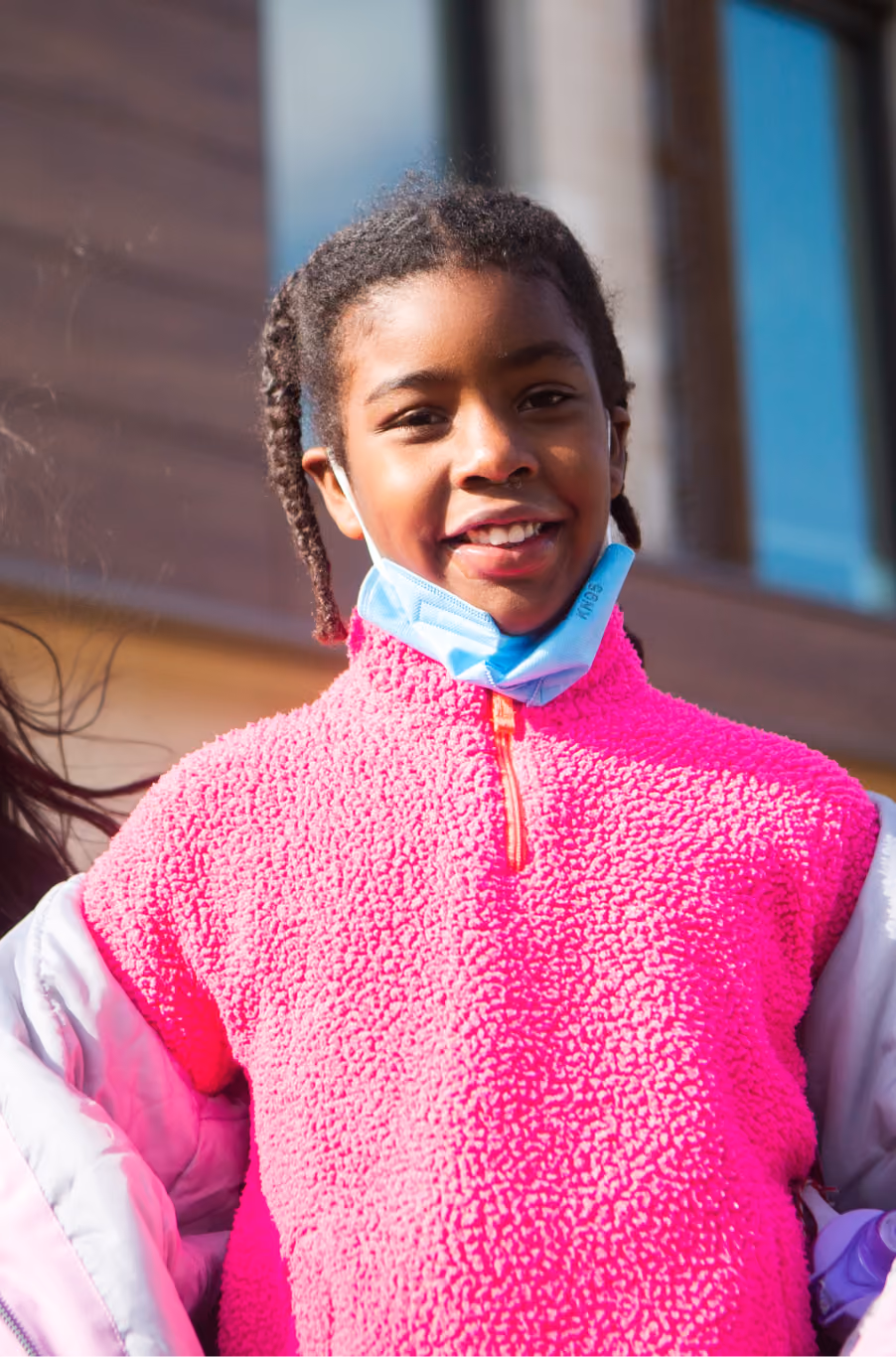 Young smiling girl with braided hair wearing a bright pink fleece jacket and a light blue face mask pulled down below her chin, standing outdoors near a building with large windows.