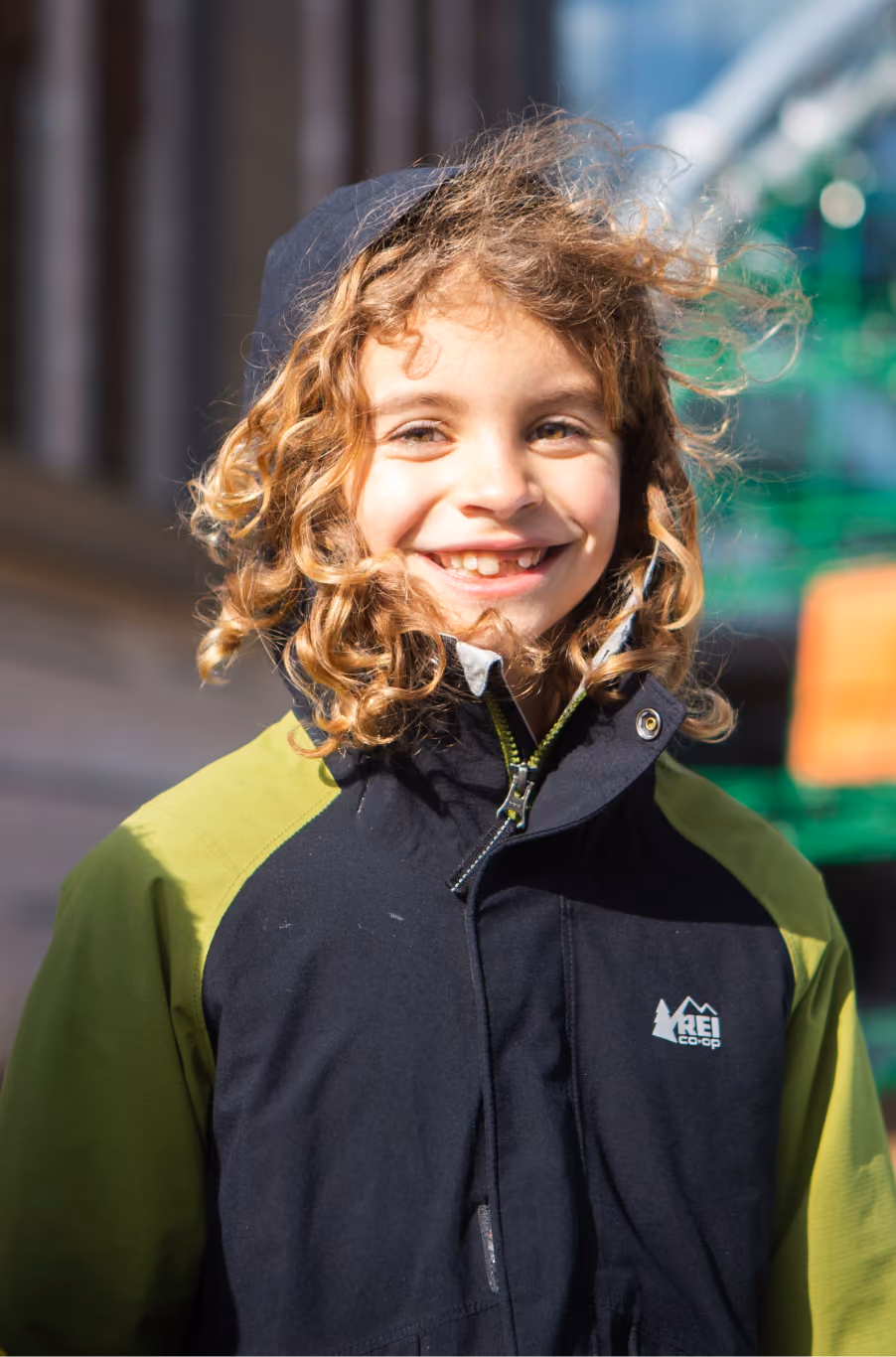 Smiling child with curly blonde hair wearing a black and green REI Co-op jacket outdoors in sunlight.