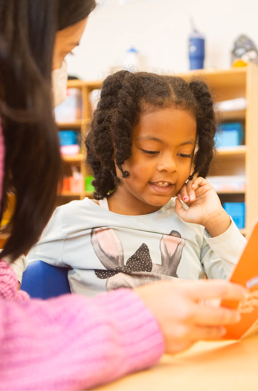 Young girl with braided hair reading a book with an adult wearing a pink sweater, in a classroom setting with shelves and colorful containers in the background.