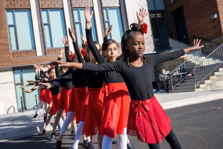 A line of young dancers outdoors wearing black tops and red skirts, raising one arm and extending the other, performing a synchronized dance in front of a building.