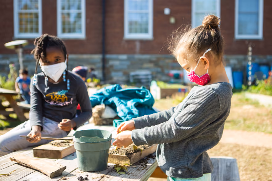 Two young children wearing face masks engage in outdoor gardening activities at a wooden table with soil and plants, against a background of a brick building and garden area.