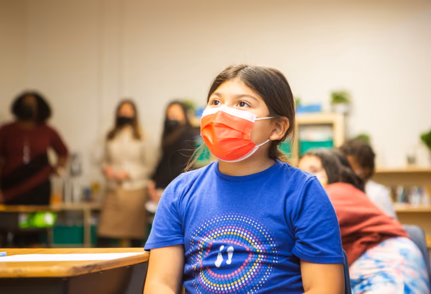 A girl wearing a blue shirt and orange face mask sits at a desk in a classroom, looking upward, with blurred adults and children in the background.