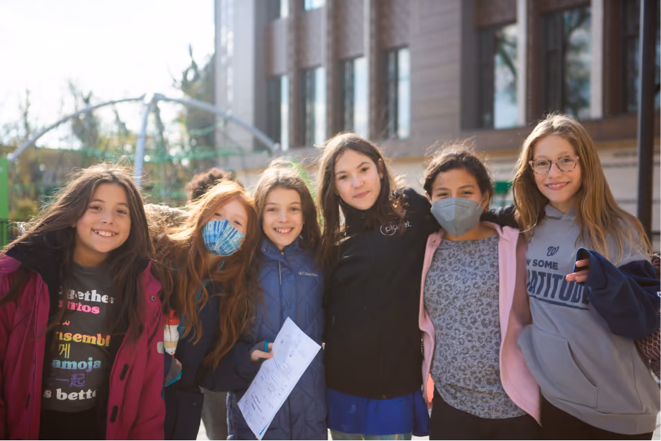 Group of six smiling young girls standing close outdoors with one holding a paper, two wearing face masks, in front of a building.