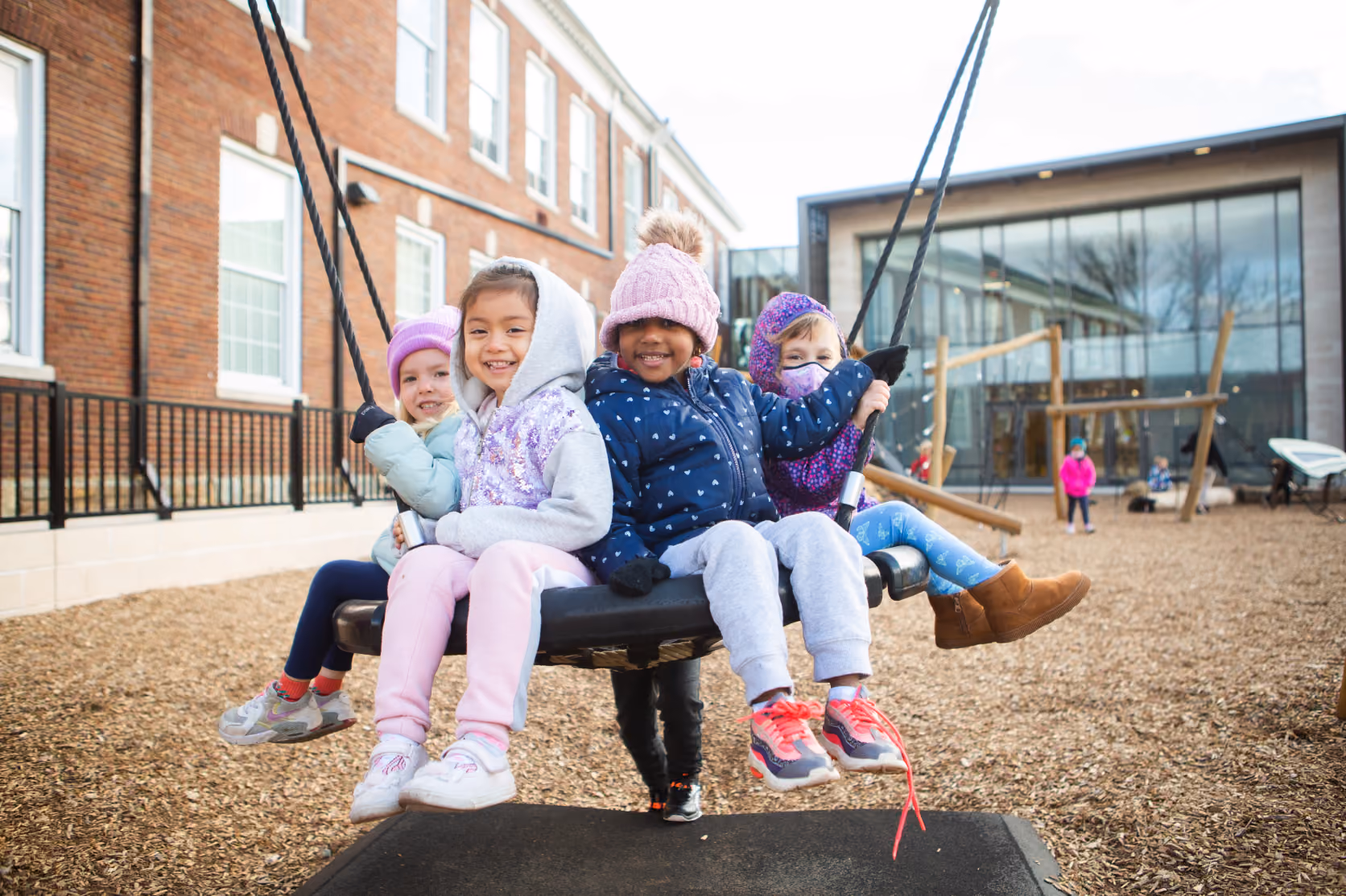 Four young children bundled in winter clothes and hats sitting together on a large round swing in a playground with a school building and other playground equipment in the background.