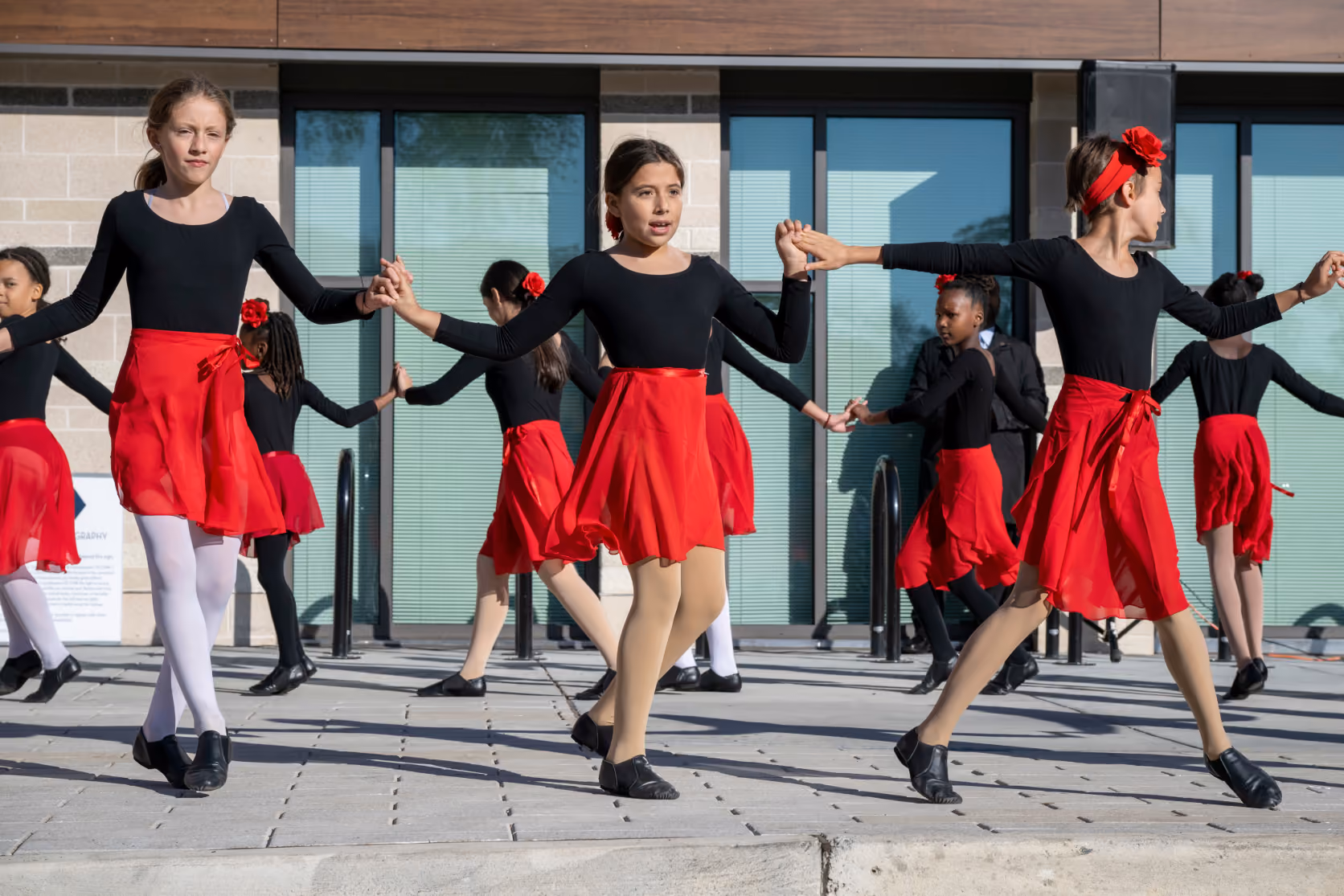 A group of young girls wearing black tops, red skirts, and black shoes perform a coordinated dance holding hands outdoors on a tiled surface in front of glass doors.