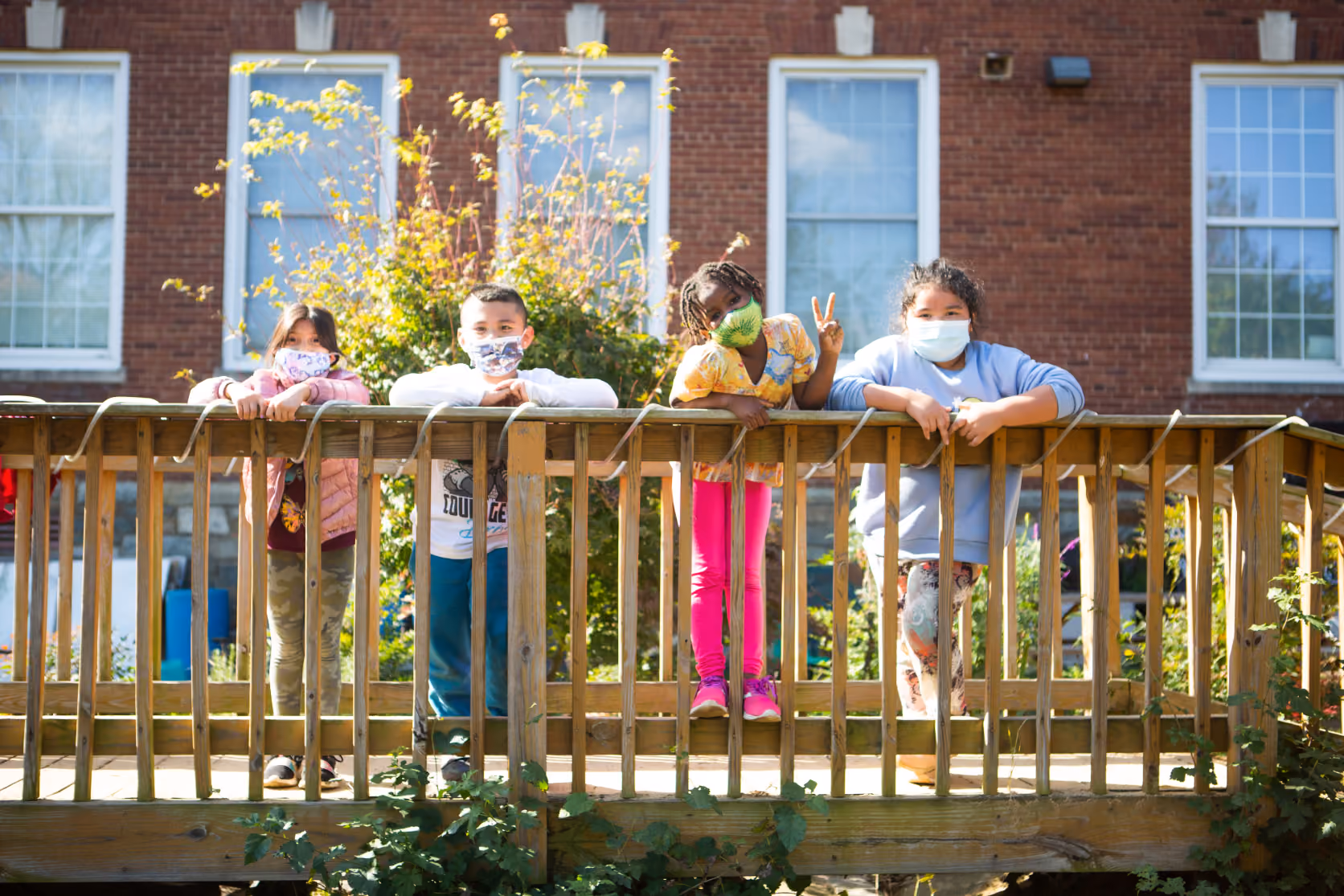 Four children wearing face masks stand leaning on a wooden railing outdoors with a brick building and windows behind them, one child making a peace sign.