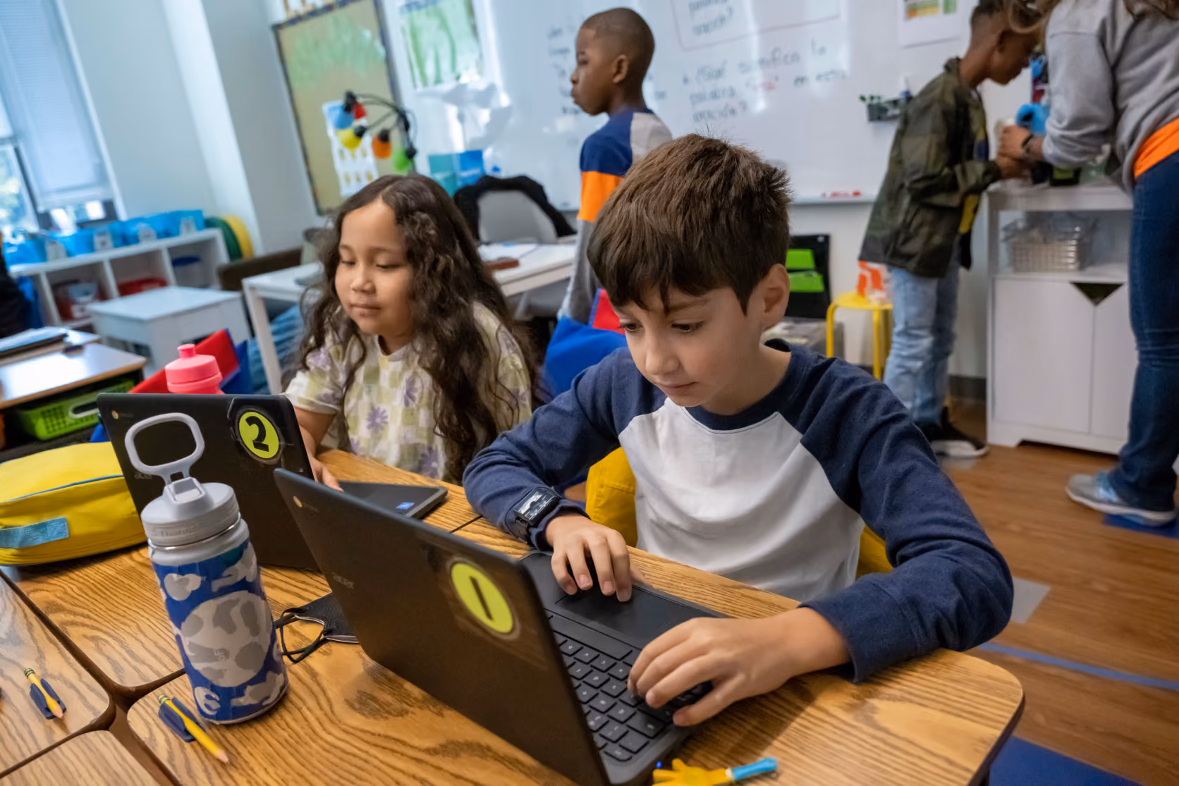 Two children sitting at a wooden classroom table using laptops with numbered stickers, a boy in a blue and white shirt focused on his laptop and a girl in a light-colored patterned shirt looking at her screen, with water bottles and school supplies nearby.