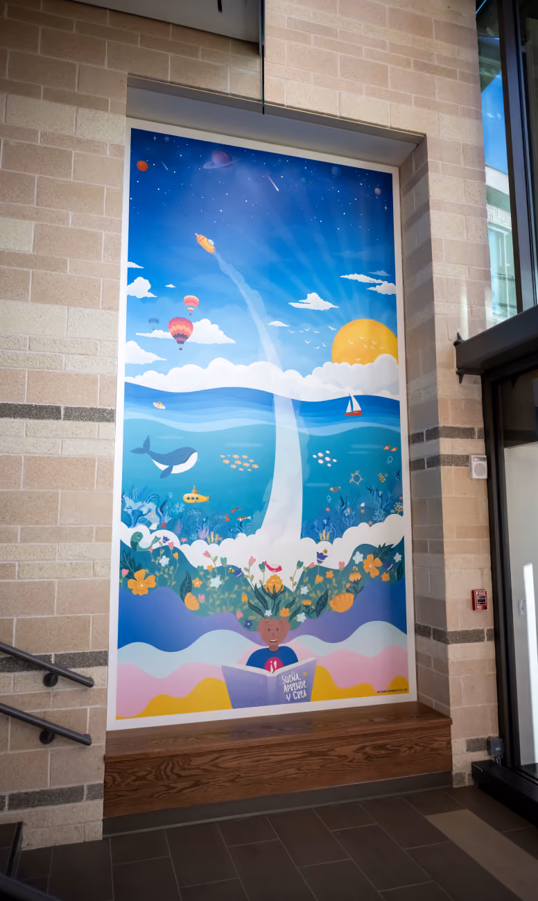 Colorful mural in an indoor alcove depicting a child reading a book titled 'CUENTO, HISTORIA Y CULA' with imaginative scenes flowing from the child's head, including ocean waves with a whale and fish, underwater plants, a yellow submarine, a sailboat on the sea, balloons and a rocket soaring into a blue sky with clouds and sun.