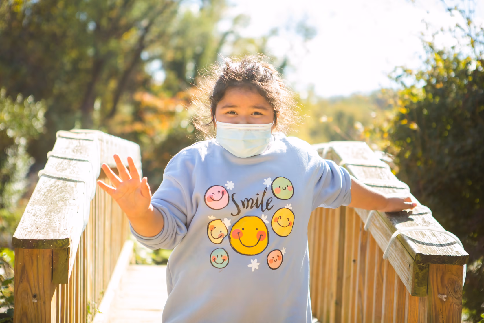 Child wearing a face mask and a light blue sweatshirt with colorful smiley faces and the word 'Smile', standing on a sunlit wooden bridge surrounded by greenery, waving with one hand.