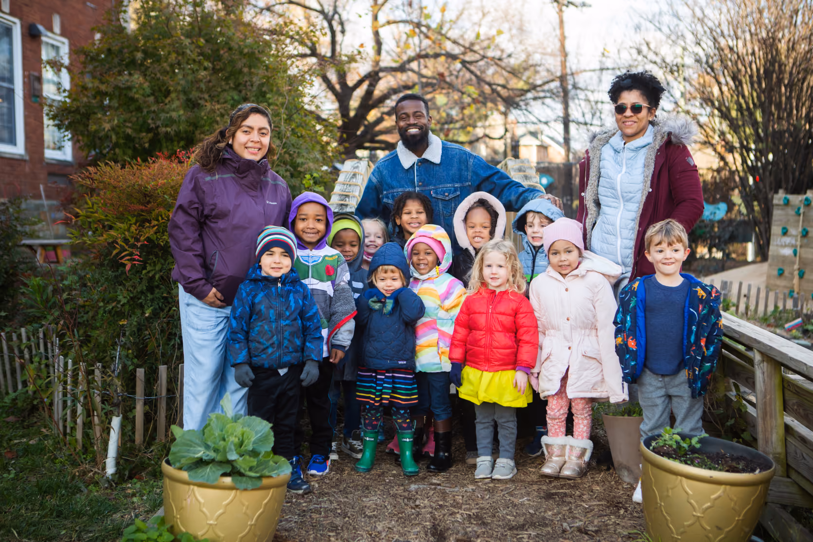 Smiling adults and children gathered outdoors in a garden area with plants, dressed in warm jackets and colorful clothing on a chilly day.